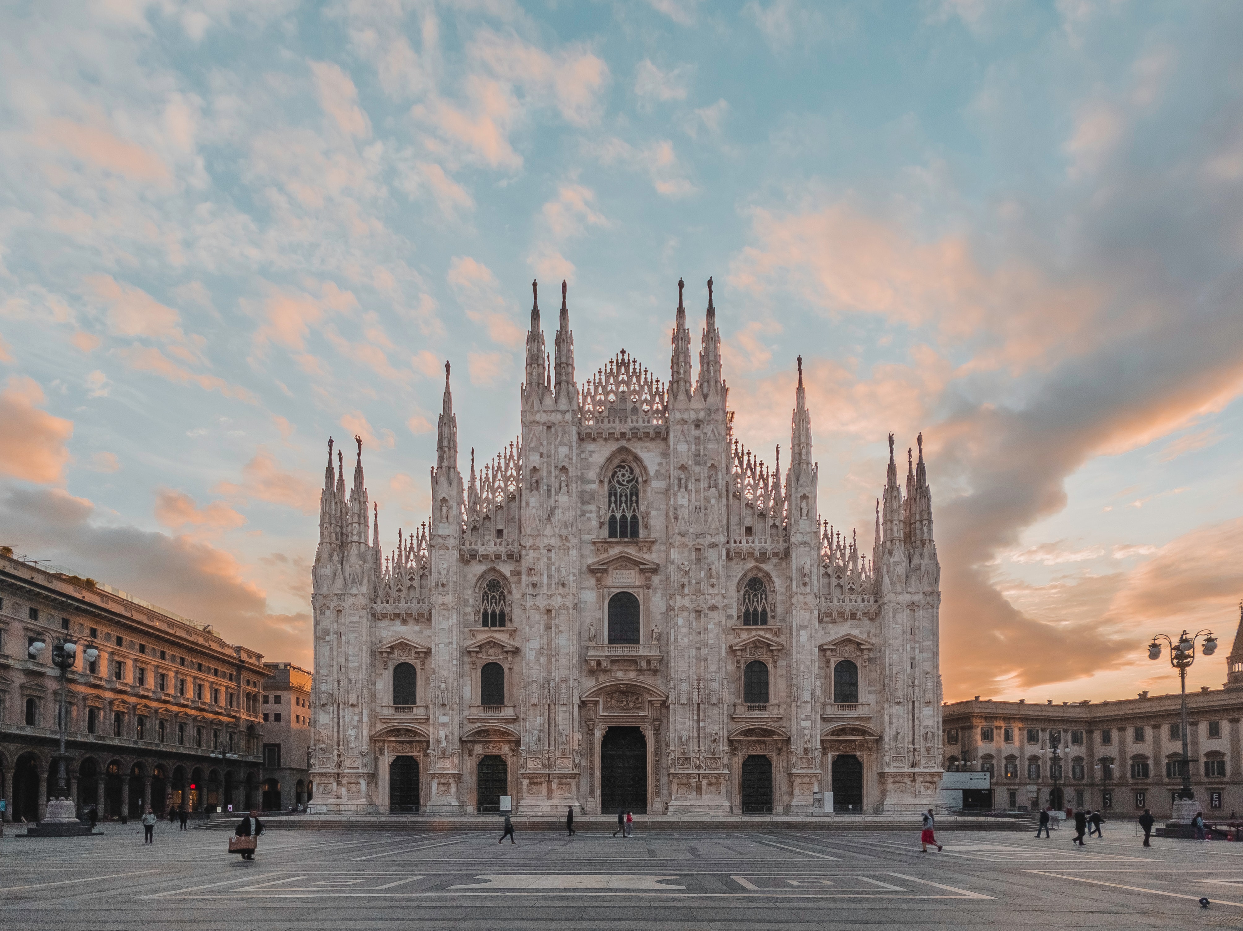 a large white building with many spires and people walking in front with Milan Cathedral in the background